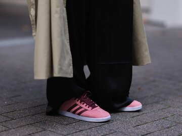 Frau mit schwarzer weiter Hose und einem beigen Trenchcoat trägt auf der Straße pinke Adidas Gazelle. | © Getty Images/Jeremy Moeller