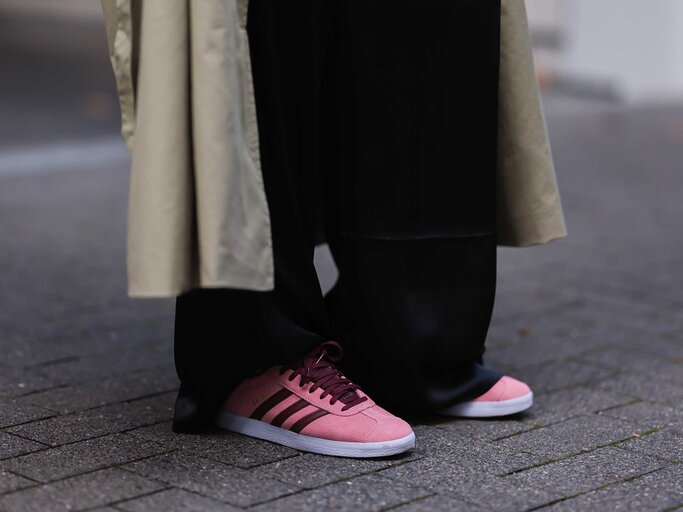 Frau mit schwarzer weiter Hose und einem beigen Trenchcoat trägt auf der Straße pinke Adidas Gazelle. | © Getty Images/Jeremy Moeller