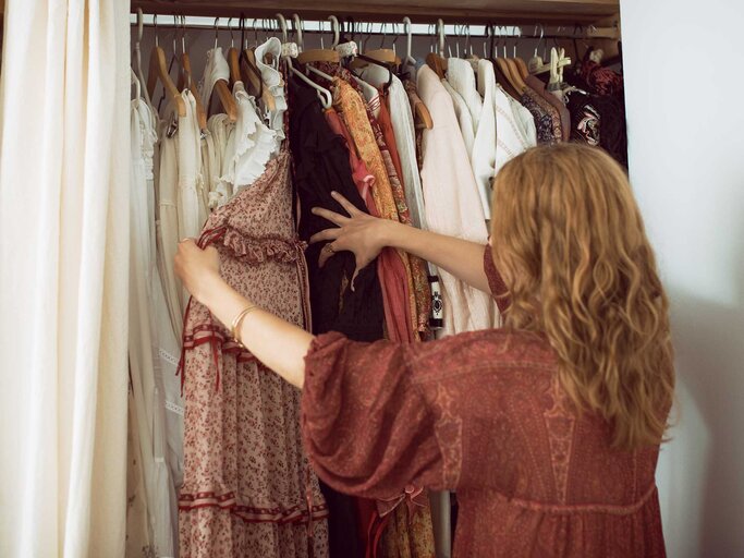 Junge Frau steht vor ihrem Kleiderschrank und hält ein Kleid in der Hand. | © Getty Images / Emilija Manevska