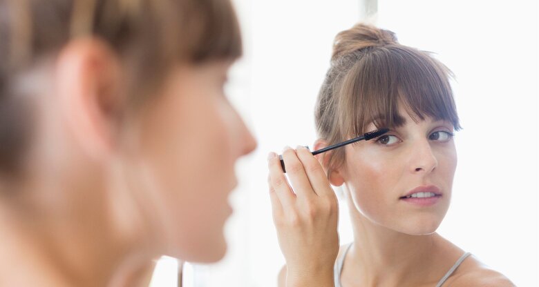 Eine Frau schaut in den Spiegel und trägt pflegendes Öl mit einer kleinen Bürste auf ihre Wimpern auf | © Getty Images / Letizia Le Fur