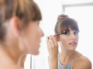 Eine Frau schaut in den Spiegel und trägt pflegendes Öl mit einer kleinen Bürste auf ihre Wimpern auf | © Getty Images / Letizia Le Fur