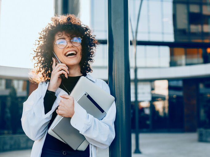 Junge Frau mit lockigen Haaren und runder Brille lacht telefonierend im Sonnenlicht, hält ein Tablet und einen Laptop. | © adobe.stock.com/Strelciuc