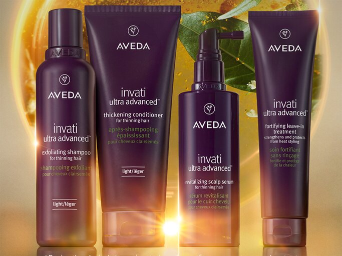 Aveda Invati Ultra Advanced Haarpflegeserie mit vier violetten Produkten vor einem Hintergrund mit goldenem Licht und grünen Blättern. | © Aveda