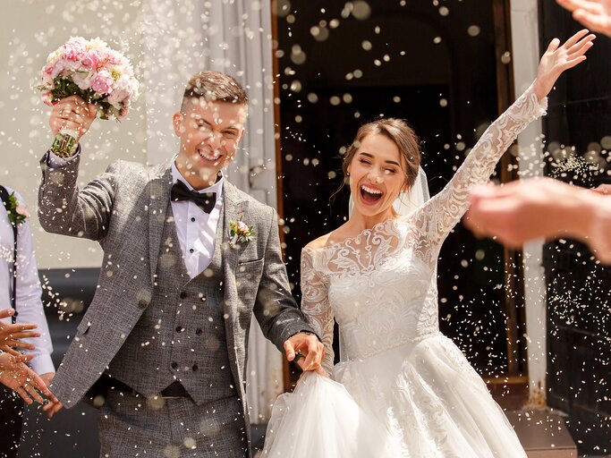 Lächelnder Bräutigam mit Blumenstrauß und Braut in weißem Kleid jubeln unter fliegendem Reis vor der Kirche. | © Wedding photography – stock.adobe.com