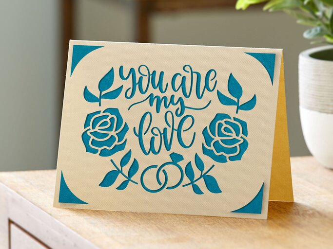 Cremefarbene Grußkarte mit blauen Rosen und dem Schriftzug "You are my love", auf einem Holztisch neben einer grünen Pflanze stehend. | © Cricut