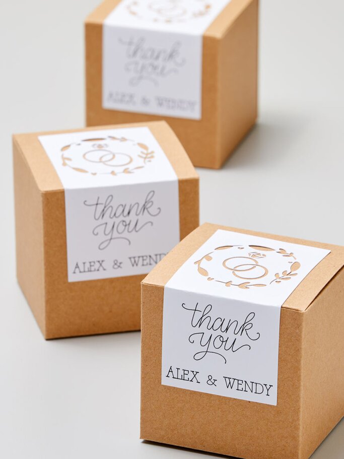 Drei braune Geschenkboxen mit weißen Etiketten, die "Thank You Alex & Wendy" und zwei Eheringe als Motiv zeigen, stehen auf einem hellen Untergrund. | © Cricut