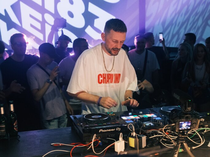 Ein DJ mit kurzem Bart und weißem T-Shirt mixt konzentriert an einem Mischpult, während eine Menschenmenge im Club ihn umgibt. | © glo