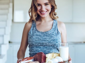 Frau in sportlichem Outfit hält ein Schneidebrett mit eiweißreichen Nahrungsmitteln wie gekochtem Ei, Kuhmilch, Käse, ein Lachssteak und rohes Fleisch | © iStock / Vasyl Dolmatov