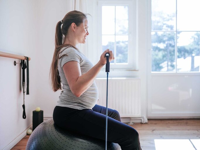 Schwangere Frau sitzt auf einem Gymnastikball mit Gummibändern in der Hand und trainiert Arme. | © Getty Images / Guido Mieth