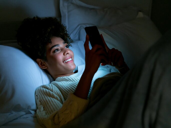 Eine Frau liegt im Bett und ist am Handy. | © GettyImages/Hispanolistic