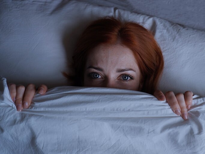 Rothaarige Frau liegt im Bett mit Bettdecke bis zur Nase gezogen und geöffneten Augen | © Getty Images/Axel Bueckert / EyeEm