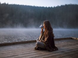 Eine Frau sitzt am frühen Wintermorgen auf einem Steg und trinkt Tee | © Getty Images / ArtistGNDphotography