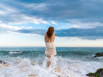 Eine Frau steht am Strand in der Brandung. Das Foto erinnert an das Gemälde "Geburt der Venus" | © Getty Images / Westend61