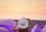 Rückansicht einer Frau in einem fließenden violetten Kleid und Hut, die inmitten blühender Lavendelfelder in den Sonnenuntergang schaut. | © Adobe Stock/MZaitsev