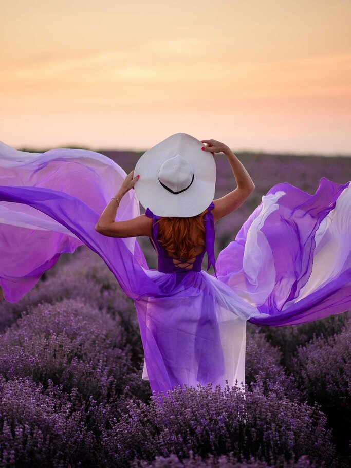 Rückansicht einer Frau in einem fließenden violetten Kleid und Hut, die inmitten blühender Lavendelfelder in den Sonnenuntergang schaut. | © Adobe Stock/MZaitsev