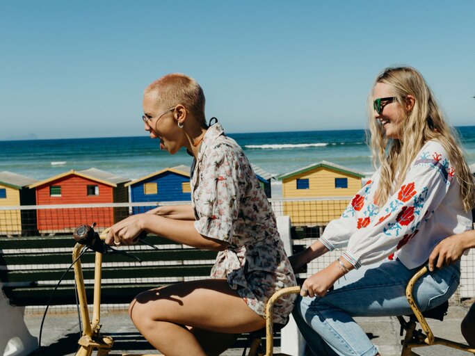 Zwei Freundinnen fahren auf einem Tandem den Strand entlang  | © Getty Images / Hello World