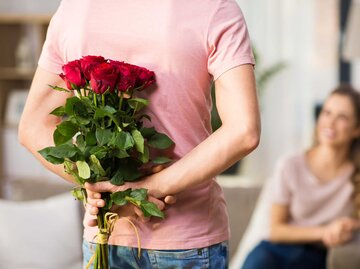 Mann hält Blumenstrauß hinter seinem Rücken, während seine Freundin auf dem Sofa sitzt | © Syda Productions