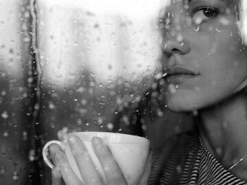 Brünette Frau sitzt an Fenster und umarmt mit traurigem Blick ihre Tasse | © iStock | yulkapopkova