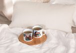 Zwei personalisierte Emaille Becher mit Fotos von einem Paar stehen auf dem Bett. | © PR