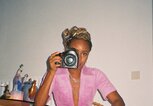 Weltfrauentag - Junge Frau sitzt im pinken Overall auf einem Hocker und hält die Kamera vor ihr Gesicht. | © PR