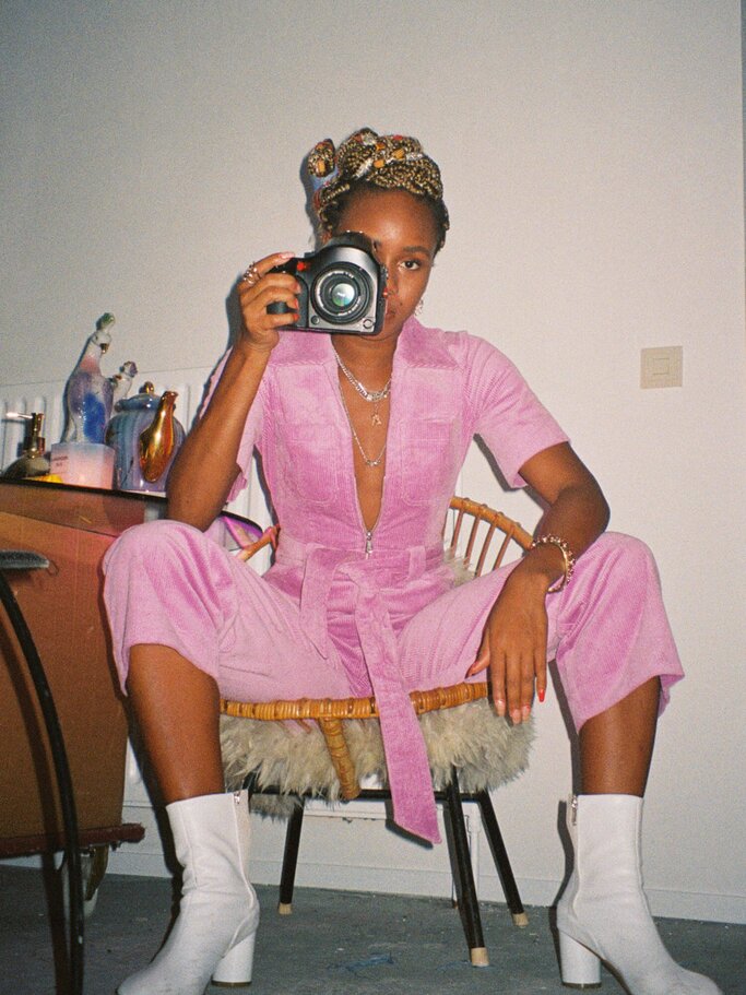Weltfrauentag - Junge Frau sitzt im pinken Overall auf einem Hocker und hält die Kamera vor ihr Gesicht. | © PR