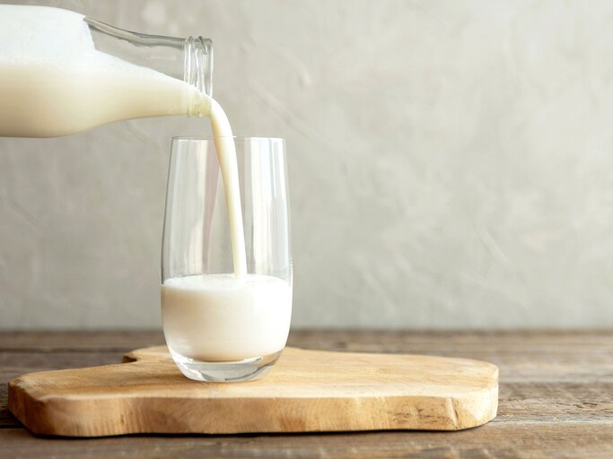 Kefir, Milch oder türkisches Ayran-Getränk werden aus einer Flasche in einen Glasbecher gegossen. Ein Glas steht auf einem Holzständer auf einem rustikalen Holztisch. | © iStock | Elena Medoks