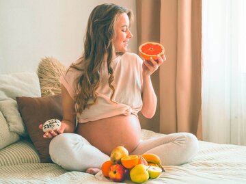 Schwangere Frau sitzt auf dem Bett mit Obst und Gemüse vor sich.  | © iStock.com / Anastasiia Stiahailo