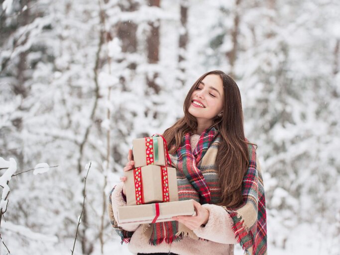 Frau steht im Schnee mit Weihnachtsgeschenken auf dem Arm.  | © iStock.com / Maya23K