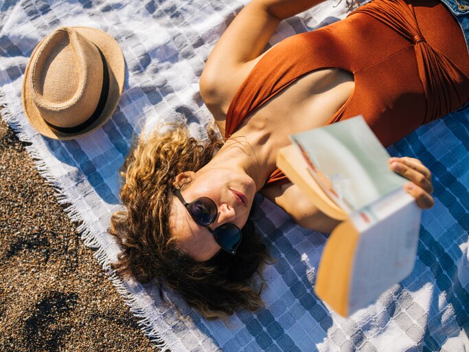 Frau liegt am Strand und liest ein Buch | © GettyImages/agrobacter