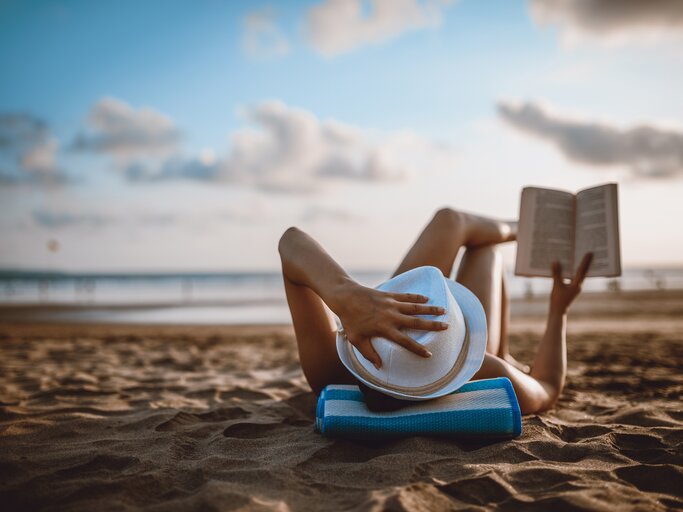 Frau liegt am Strand und liest ein Buch | © GettyImages/AleksandarGeorgiev