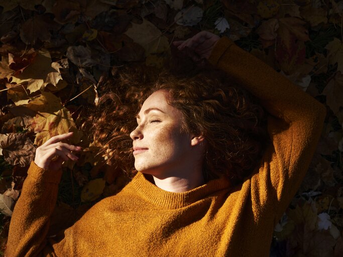 Schöne Frau liegt im Herbstlaub, während ihr Herbstsonne ins Gesicht scheint | © GettyImages/Westend61