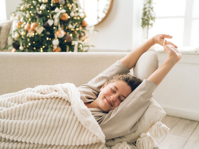 Frau liegt entspannt unter einer Decke auf dem Sofa und streckt sich, im Hintergrund steht Weihnachtsbaum | © GettyImages/Anna Efetova