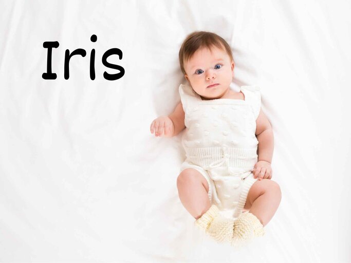 Ein Baby mit beigen Strickoverall und -söckchen liegt auf einem Bett, oben links steht der Mädchennamen Iris. | © Adobe Stock/uv_group