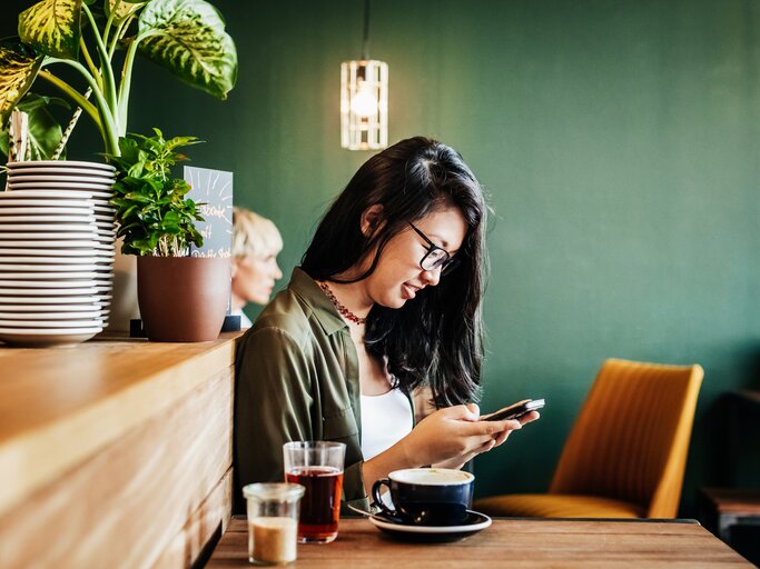 Junge Frau sitzt in einem Café und checkt ihr Smartphone | © Getty Images/	Tom Werner